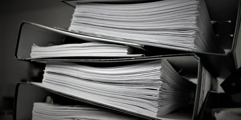 Ring binders stacked on a desk, each one overfilled with loose paper spilling past the covers.